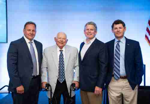 Four men in suits smile standing in front of a blue background